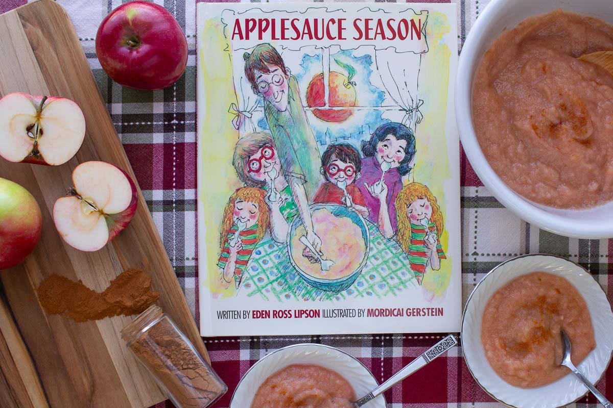 The cover of the book Applesauce Season surrounded by bowls of homemade appleauce and apples on a cutting board.