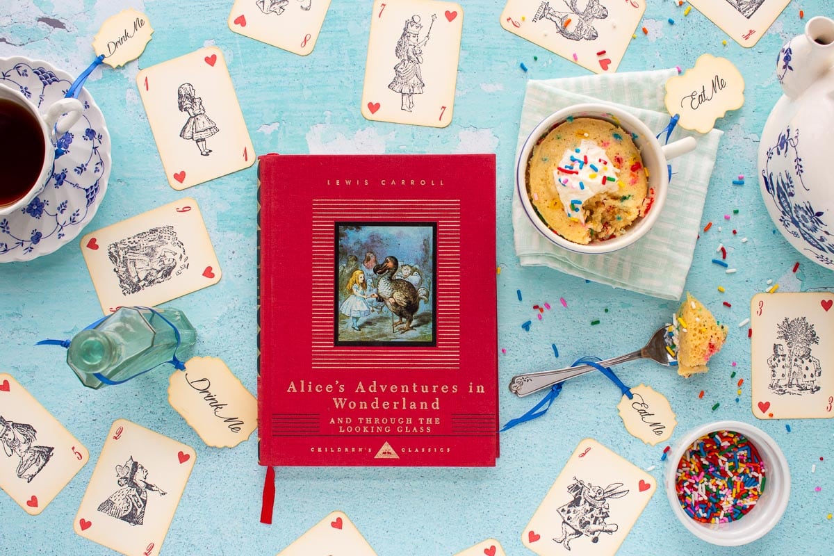 The cover of the book Alice's Adventures in Wonderland next to an Unbirthday Mug Cake with a bite taken out of it. Alice-themed playing cards are scattered on the surface.