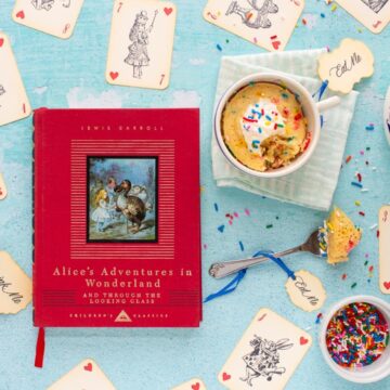 The cover of the book Alice's Adventures in Wonderland next to an Unbirthday Mug Cake with a bite taken out of it. Alice-themed playing cards are scattered on the surface.