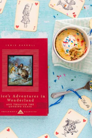 The cover of the book Alice's Adventures in Wonderland next to an Unbirthday Mug Cake with a bite taken out of it. Alice-themed playing cards are scattered on the surface.