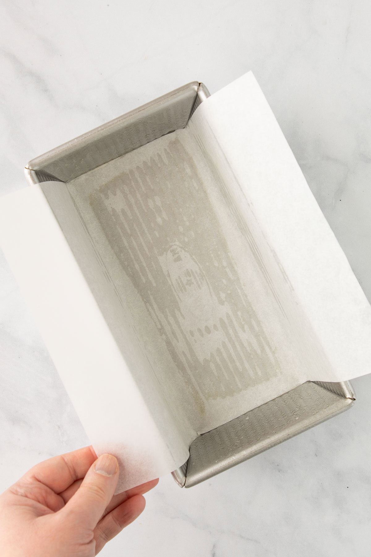 A hand lining a loaf pan with parchment paper.
