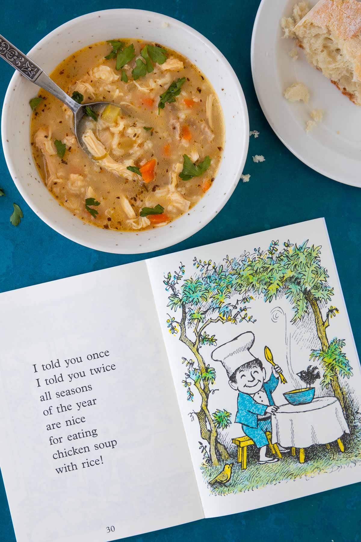The book Chicken Soup with Rice open to a page, next to a bowl of chicken soup and a piece of bread.