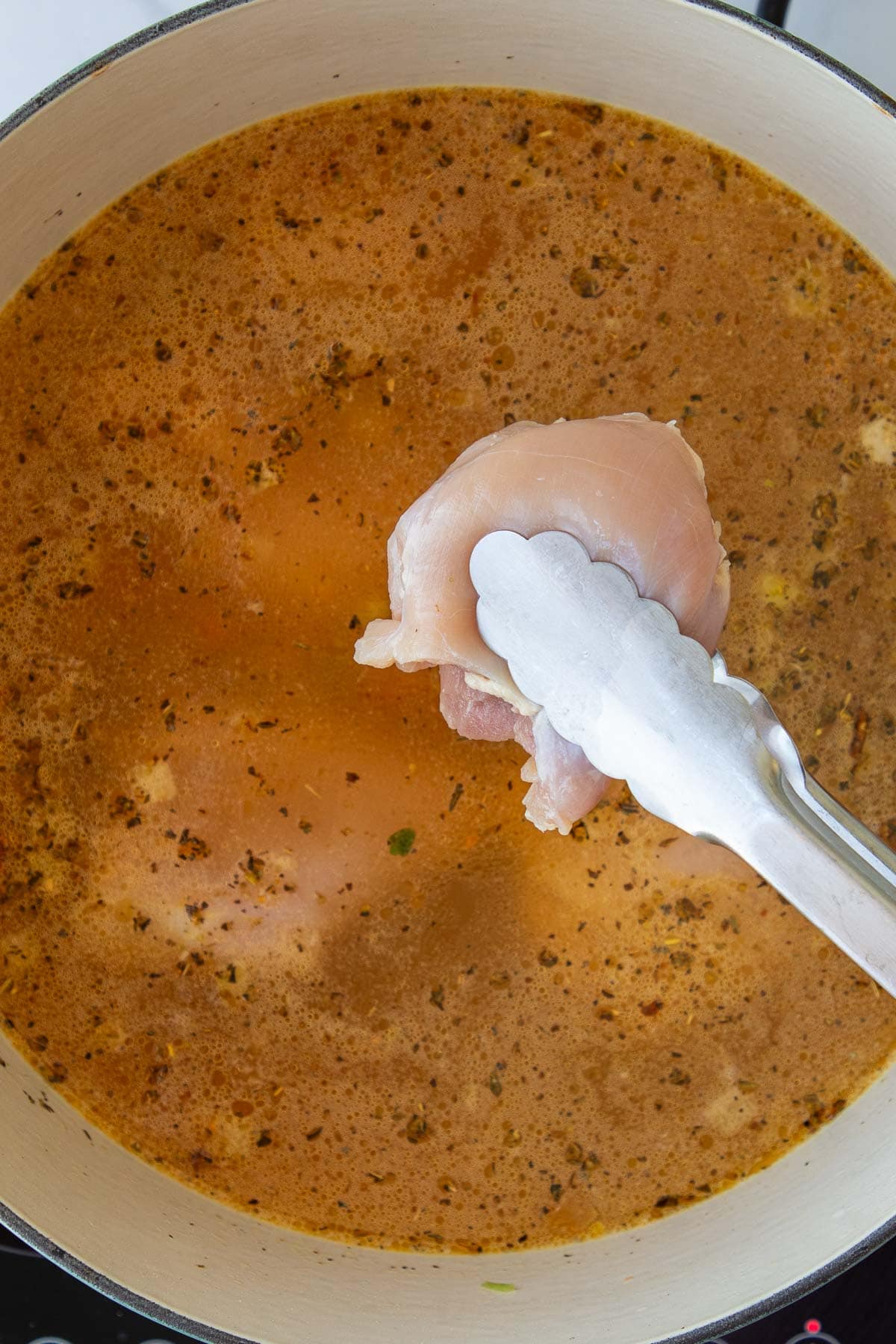 A pair of tongs lowering a chicken thigh into a pot of chicken soup.