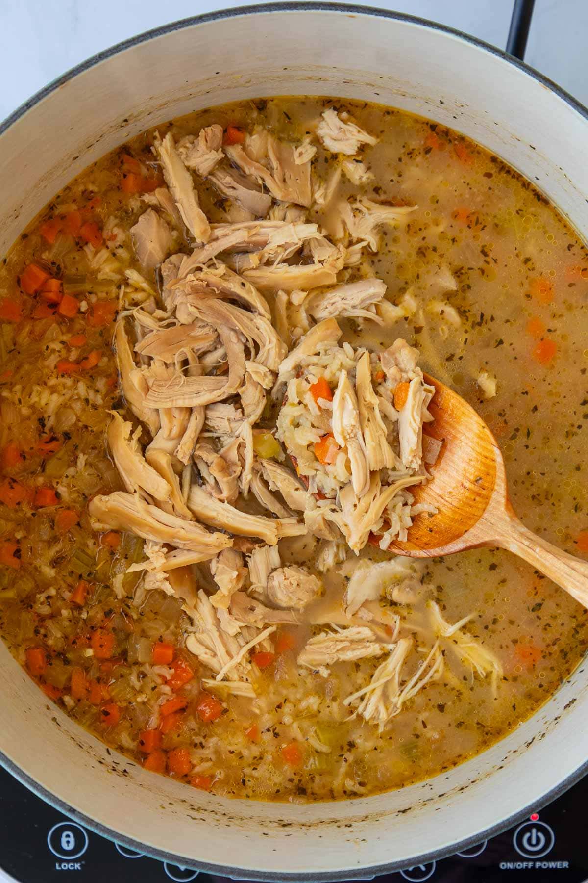 A spoon stirring shredded chicken into a pot of chicken soup with rice.