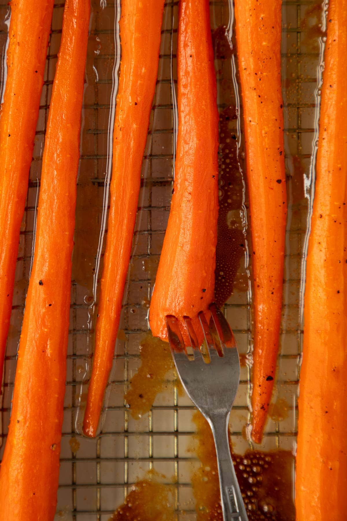 A fork poking into tender honey roasted carrots in a baking dish set on a cooling rack.