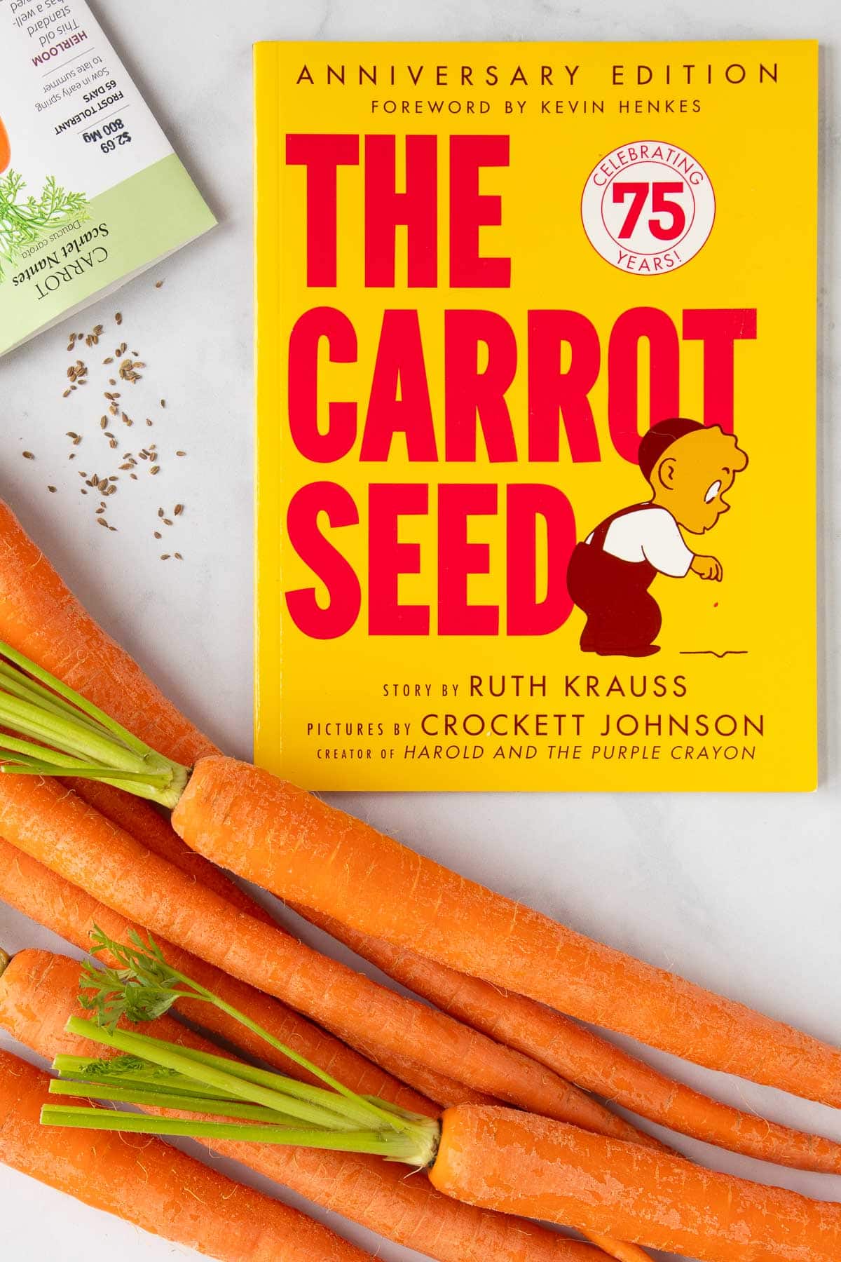 The cover of the book The Carrot Seed next to a bunch of carrots and a packet of carrot seeds.