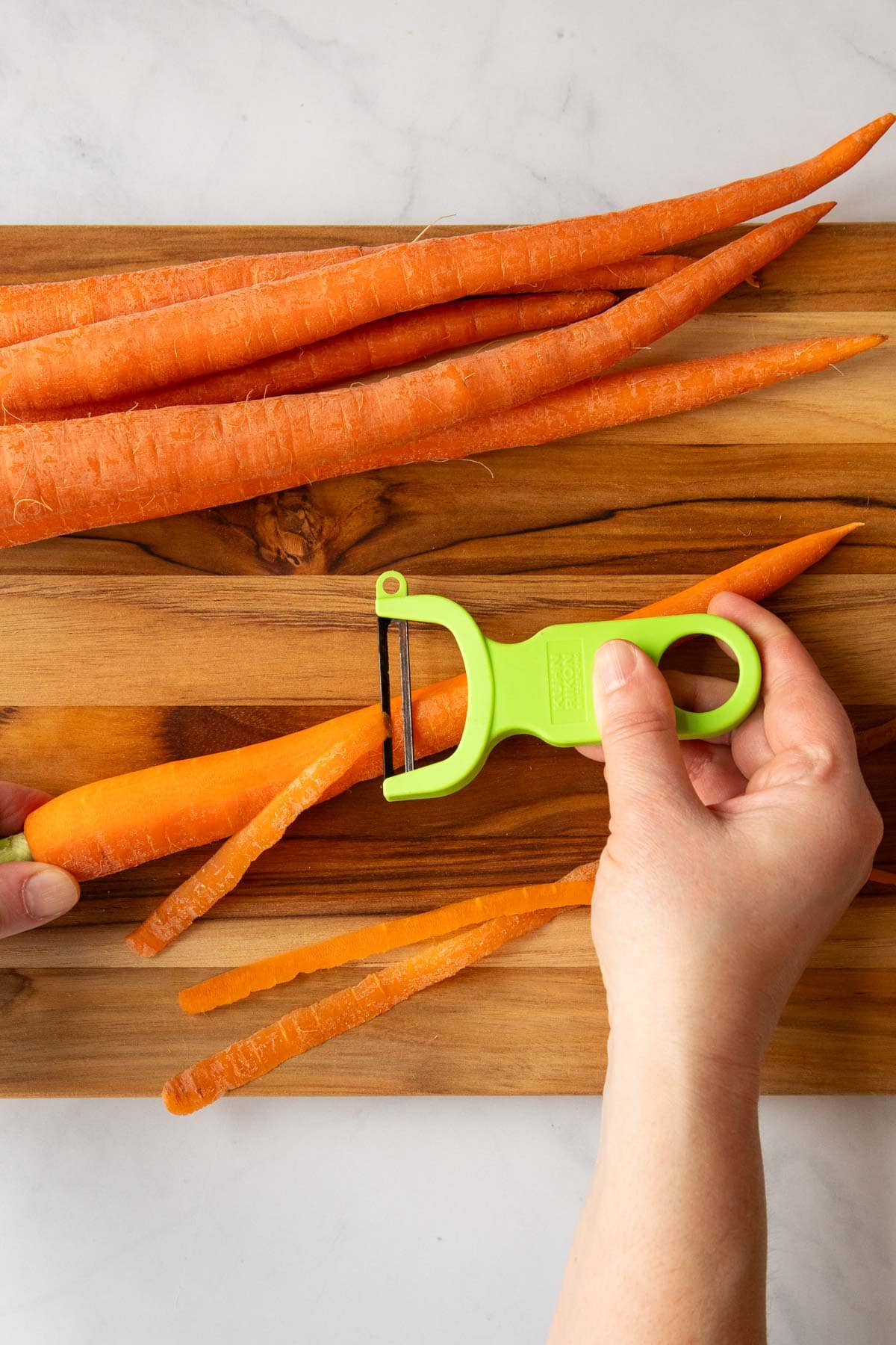 A hand using a vegetable peeler to peel a carrot on a cutting board.