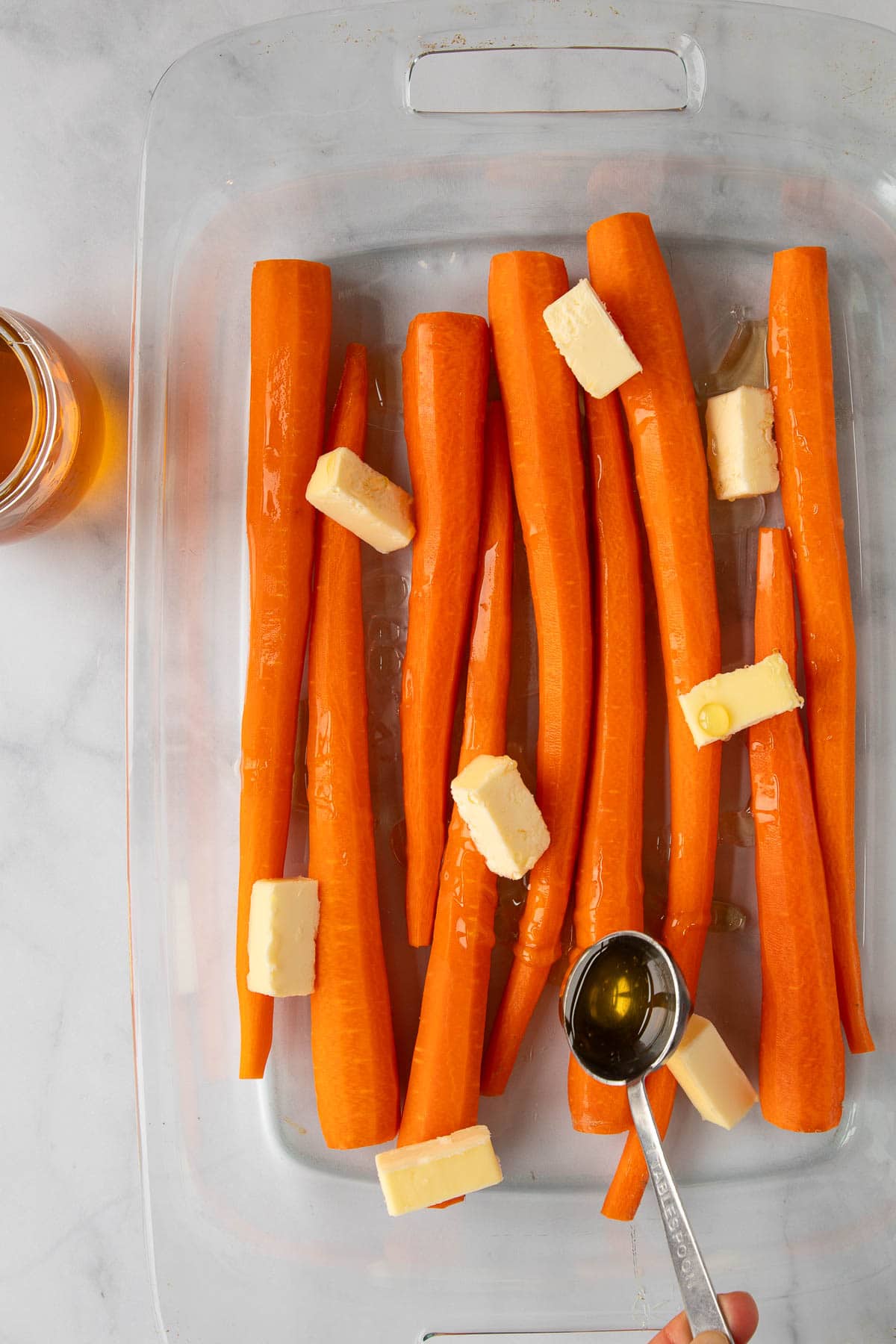 A hand drizzling honey from a tablespoon over peeled carrots and butter pieces in a baking dish.
