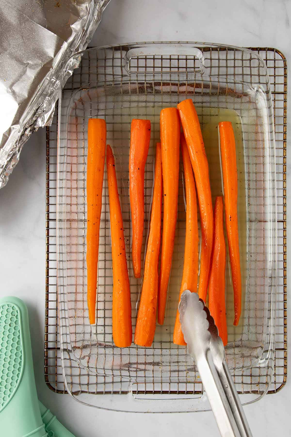 Tongs turning cooked honey roasted carrots in a baking dish set on a cooling rack.