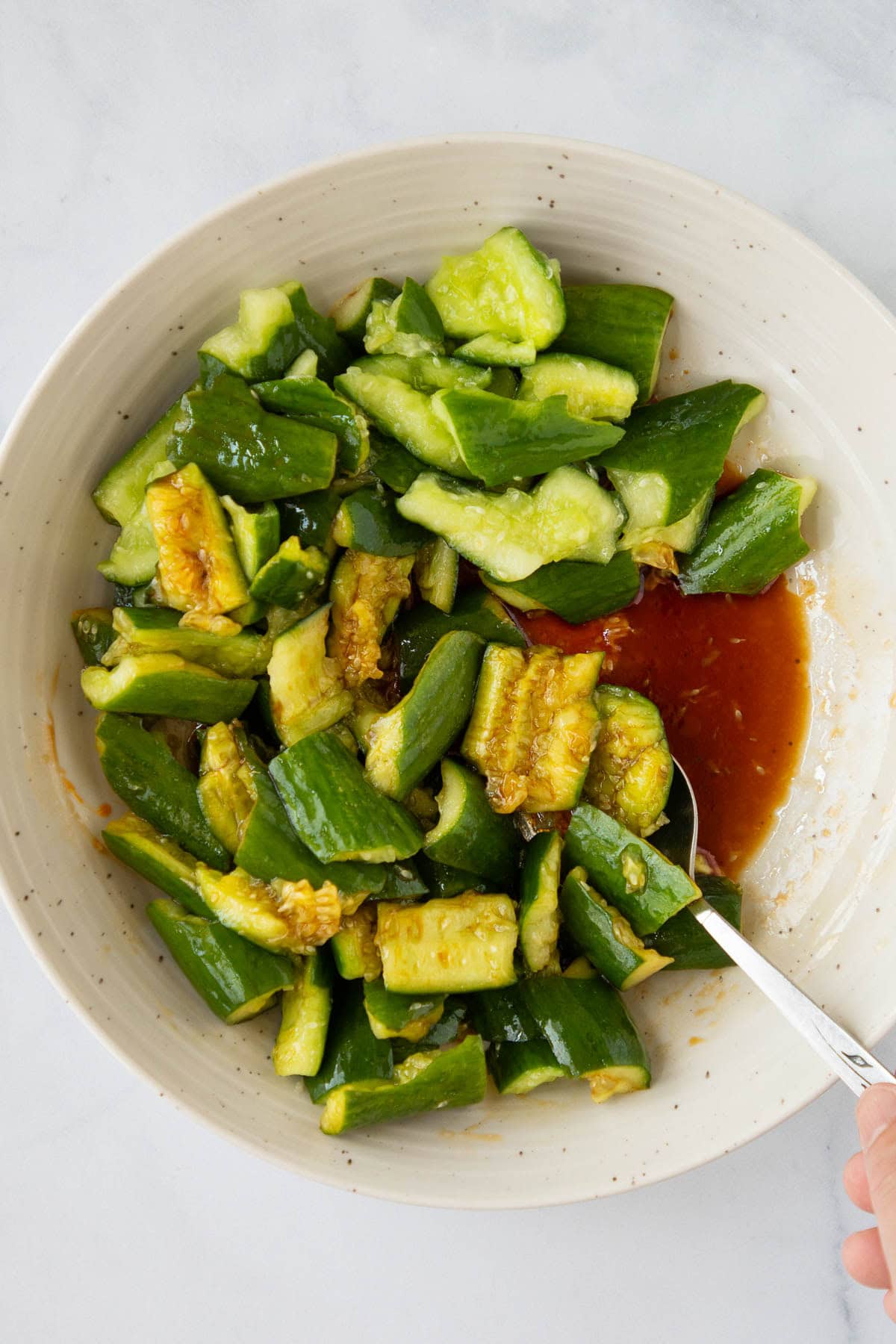 A hand using a spoon to stir smashed cucumbers in a bowl of dressing.