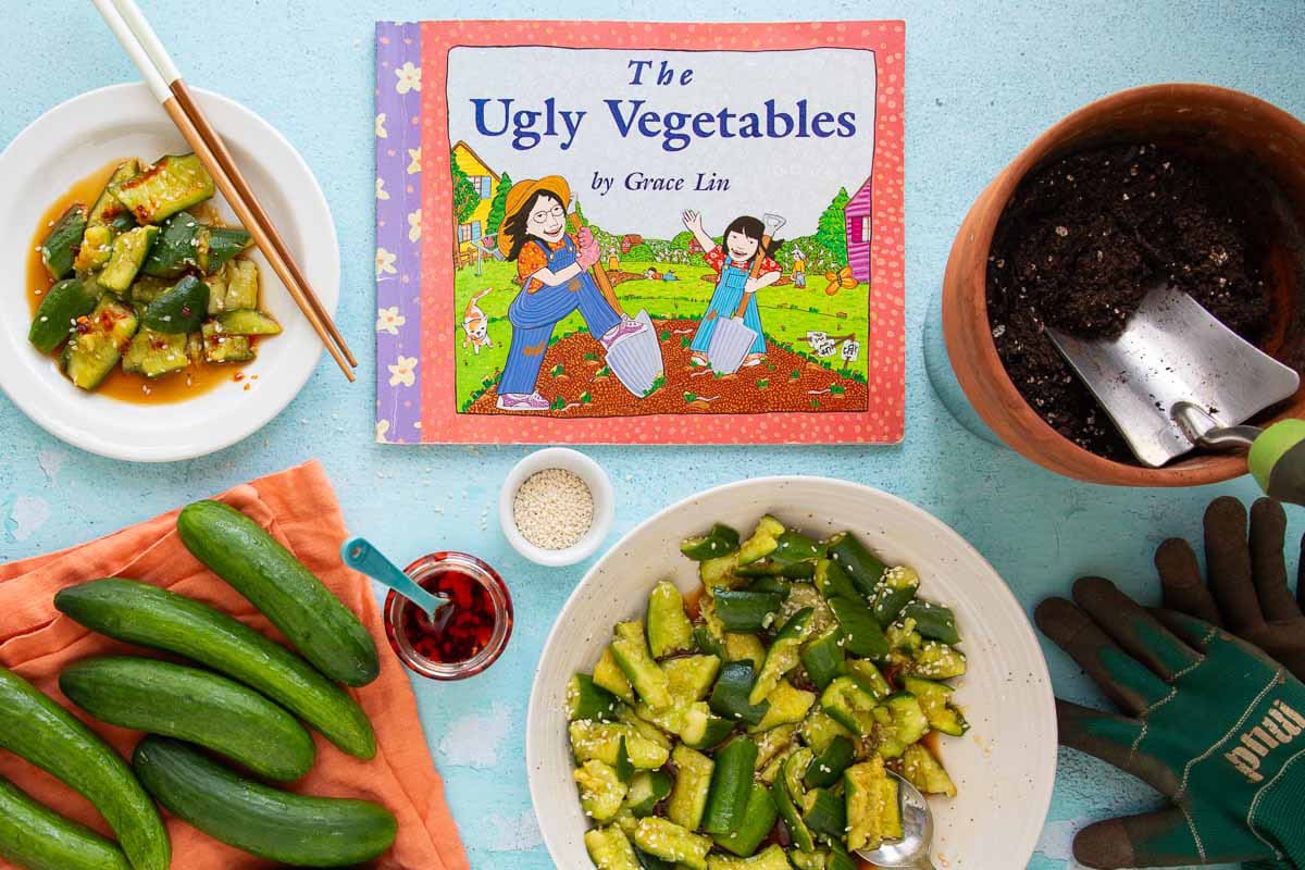 The cover of the book The Ugly Vegetables, with a bowl and plate of smashed cucumber salad, a jar of chili crisp, a pot with soil, a pair of gardening gloves, and a pile of cucumbers nearby.