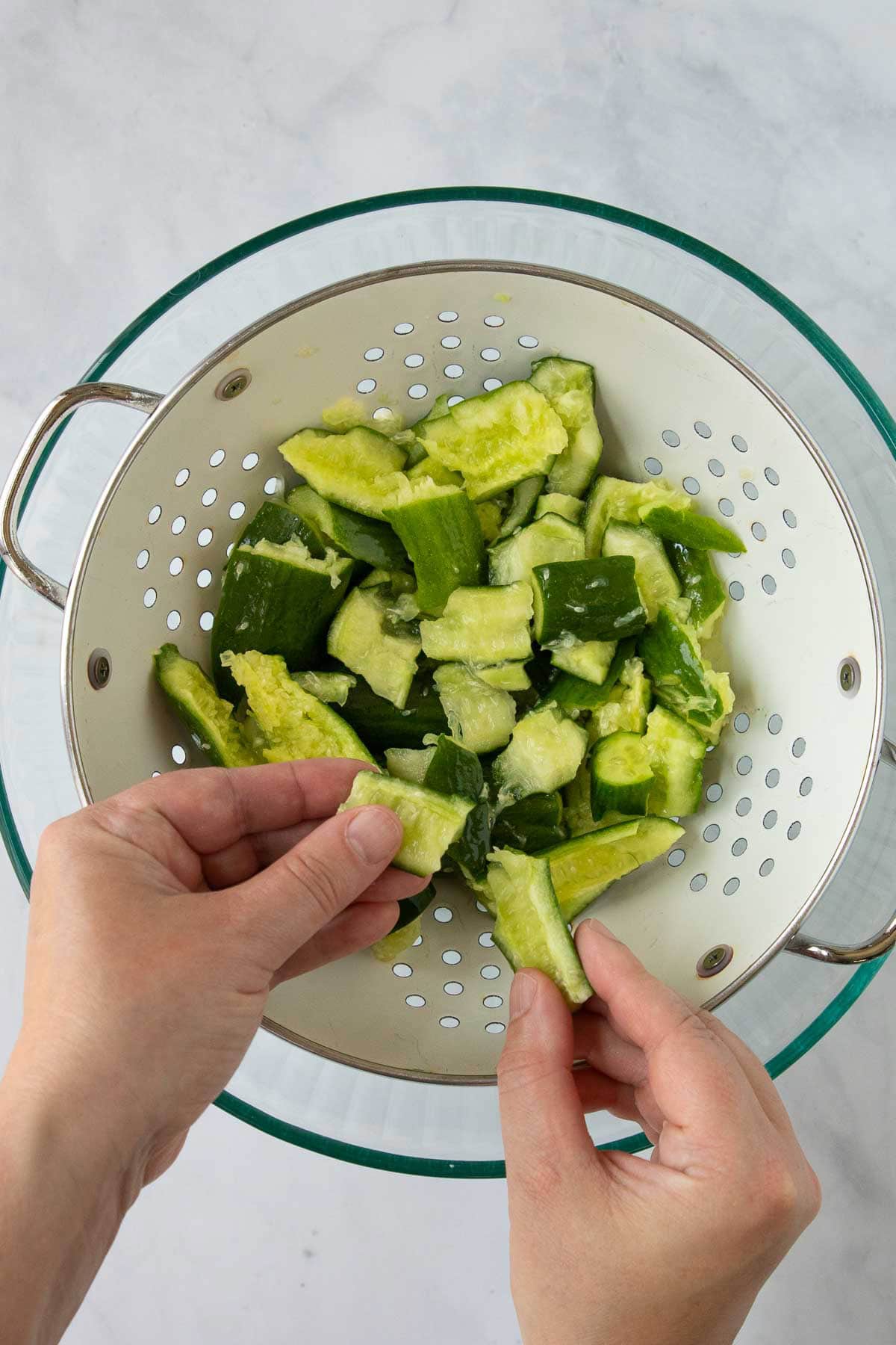 Hands tearing smashed cucumbers into pieces over a colander.