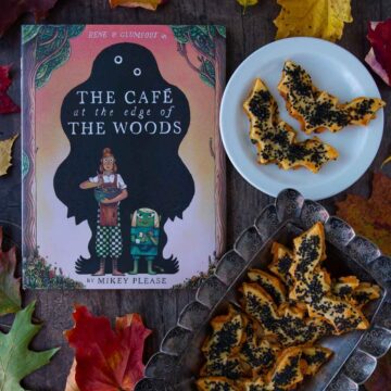 The cover of the book The Cafe at the Edge of the Woods next to a plate and tray full of cheddar-artichoke bat tarts, surrounded by colorful fall leaves.