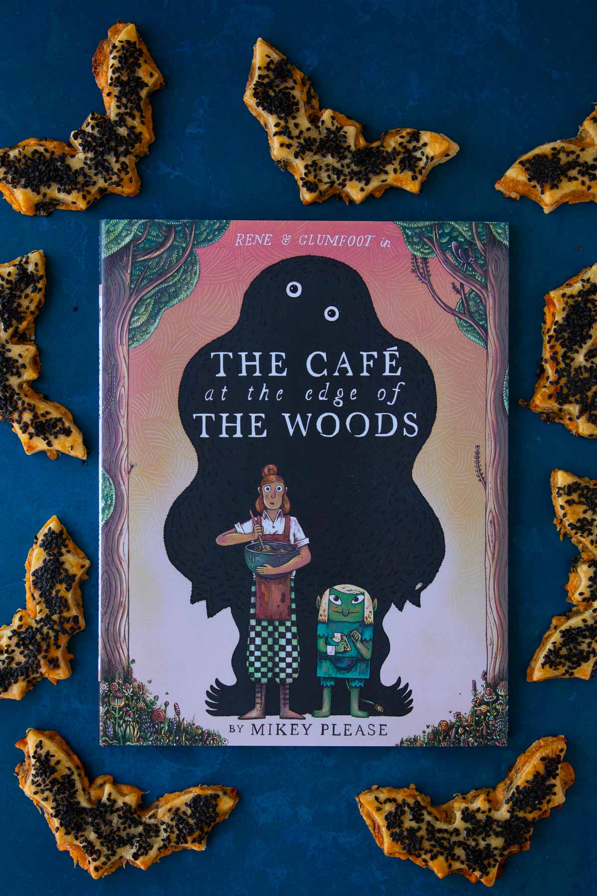 The cover of the book The Cafe at the Edge of the Woods on a blue surface, surrounded by cheddar-artichoke bat tarts.