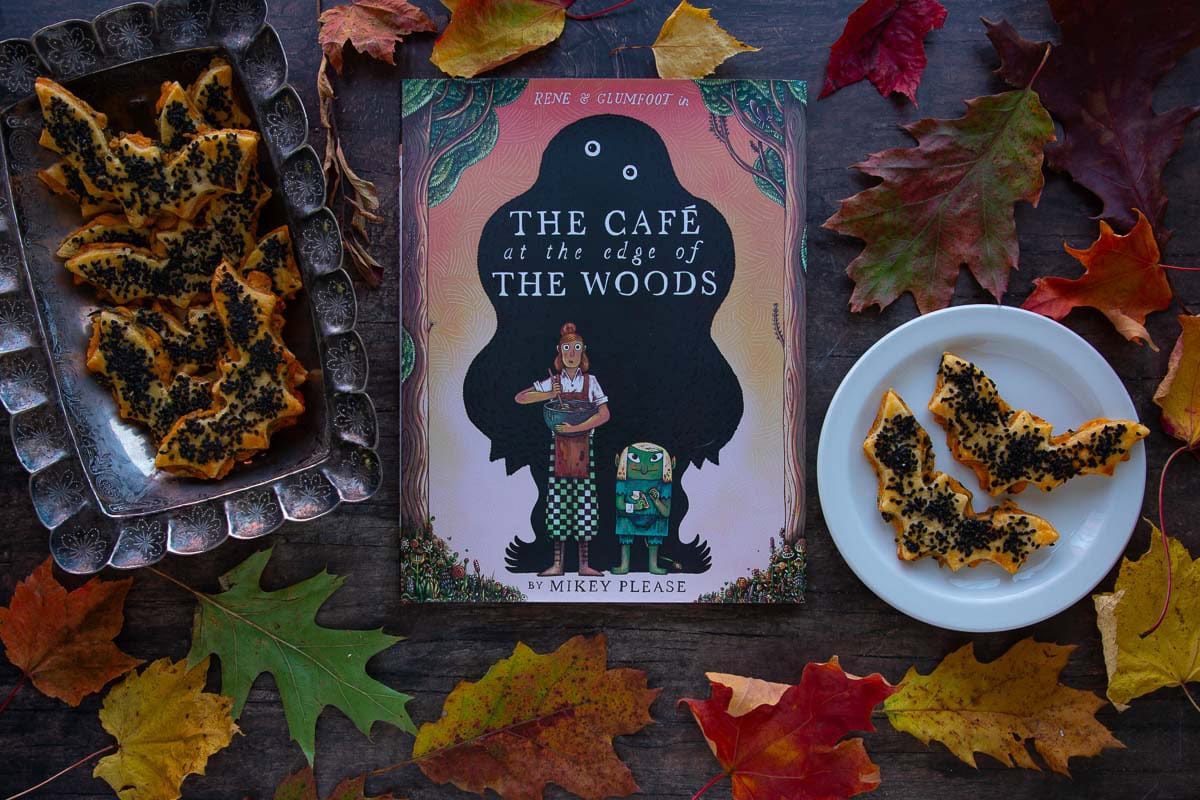The cover of the book The Cafe at the Edge of the Woods next to a plate and tray full of cheddar-artichoke bat tarts, surrounded by colorful fall leaves.