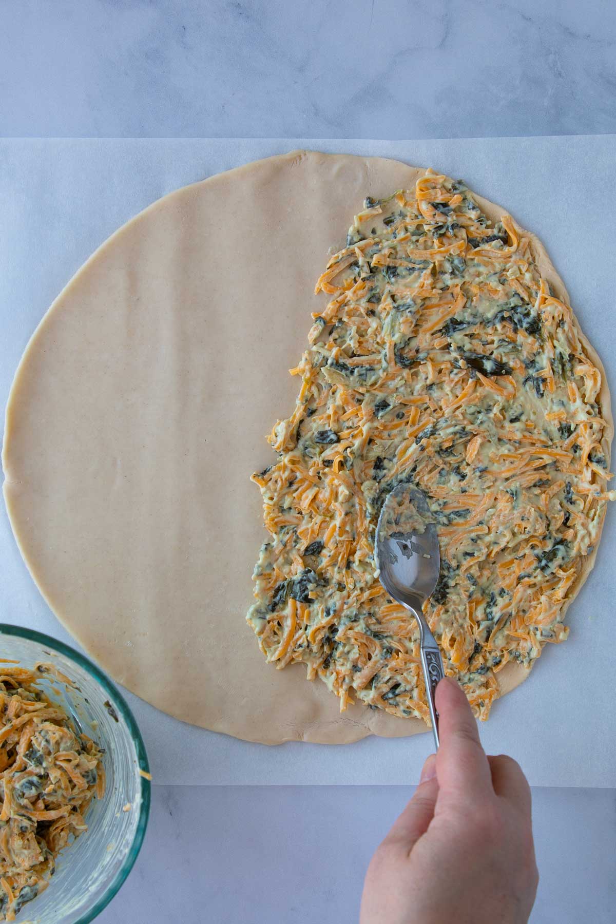 A hand using a spoon to spread out cheddar and artichoke filling on pie dough.