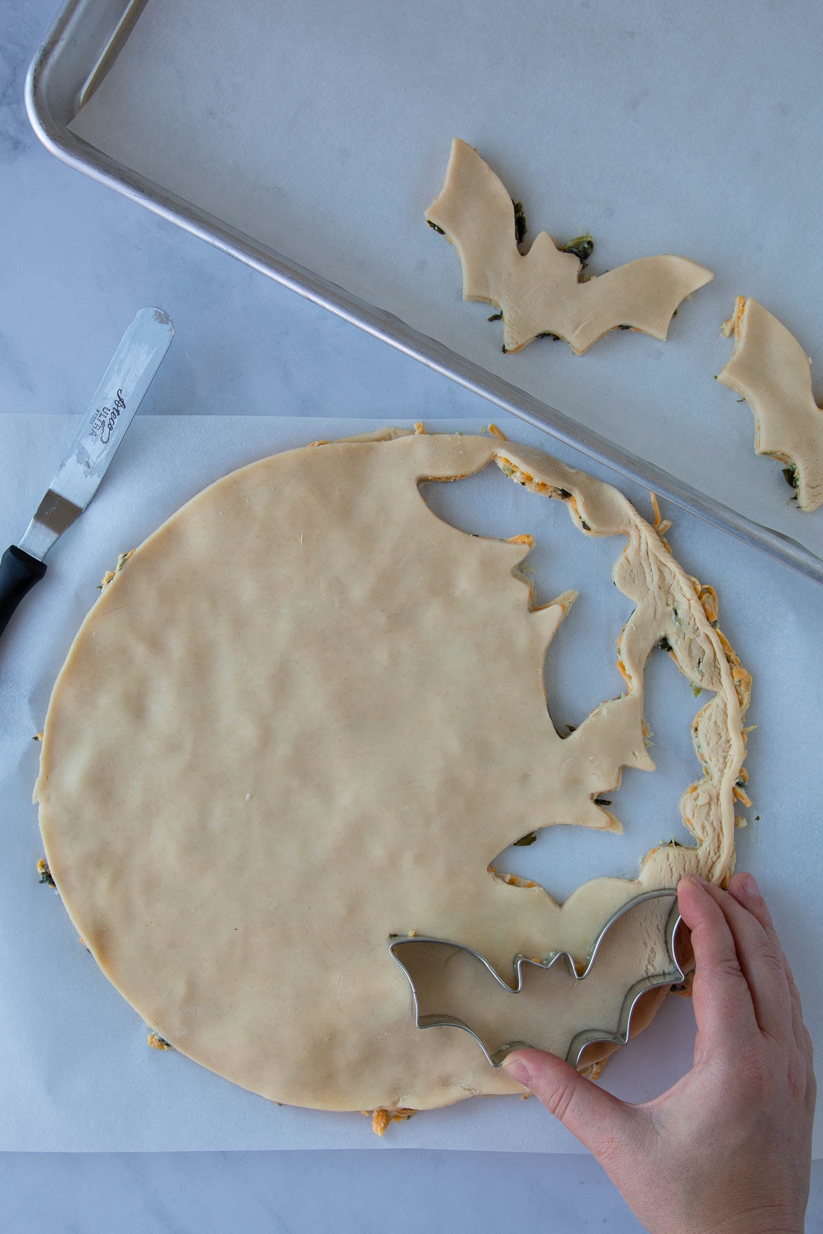A hand using a bat-shaped cookie cutter to cut out cheddar-artichoke bat tarts from pie dough.