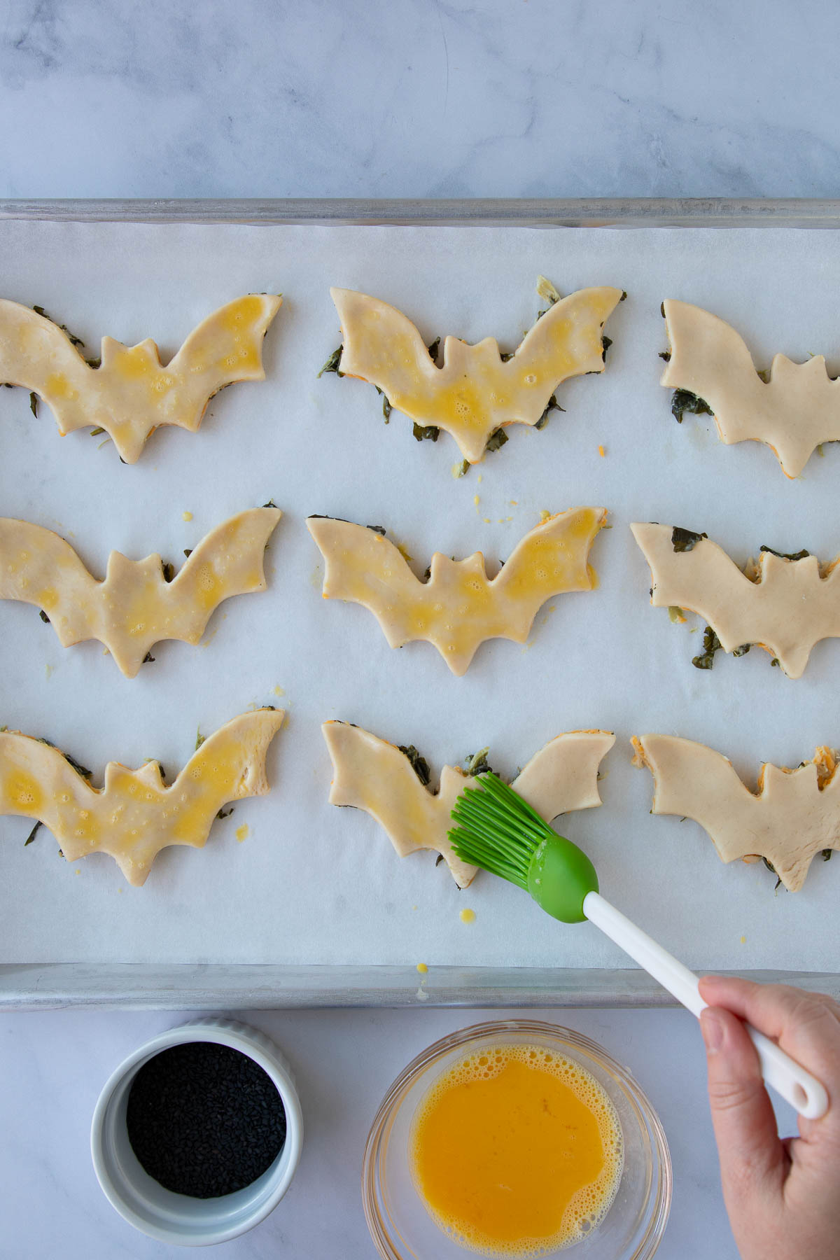 A hand using a pastry brush to paint unbaked cheddar-artichoke bat tarts with egg wash.